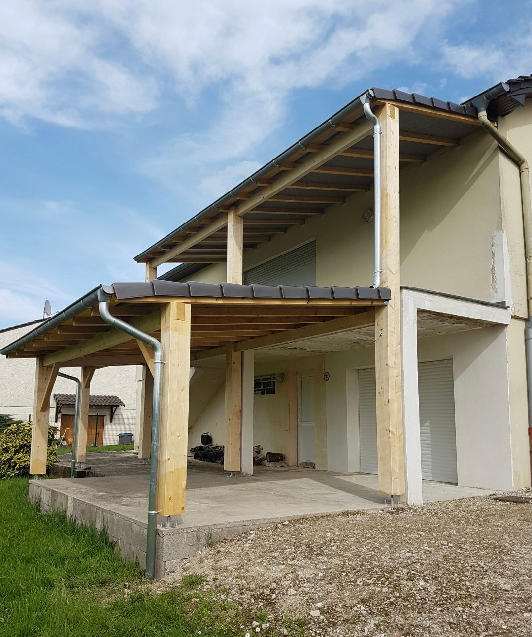 Pergola en bois en construction, adossée à une maison beige de deux étages ; tuiles grises, base en béton.