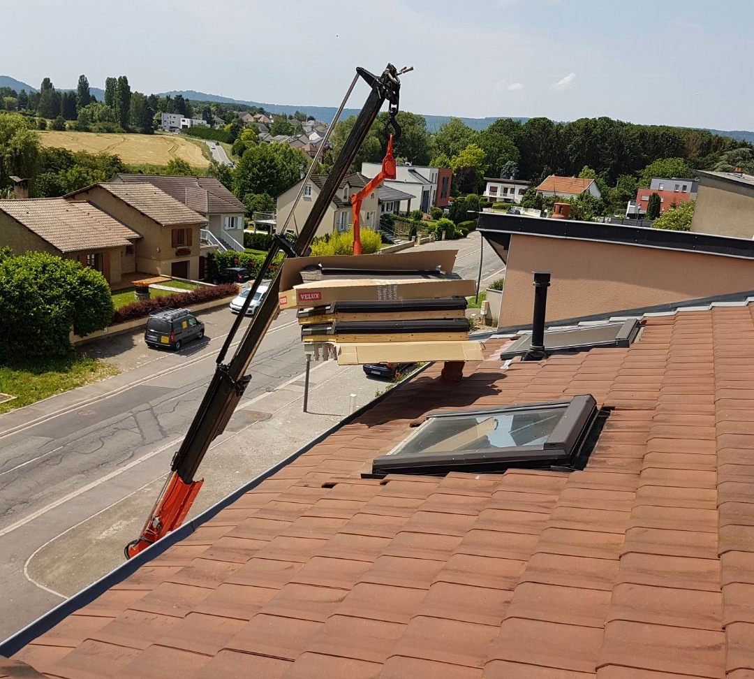 Une grue soulève des matériaux de construction et les dépose sur un toit de tuiles rouges percé d'une lucarne ; des maisons et une rue se dessinent à l'arrière-plan.
