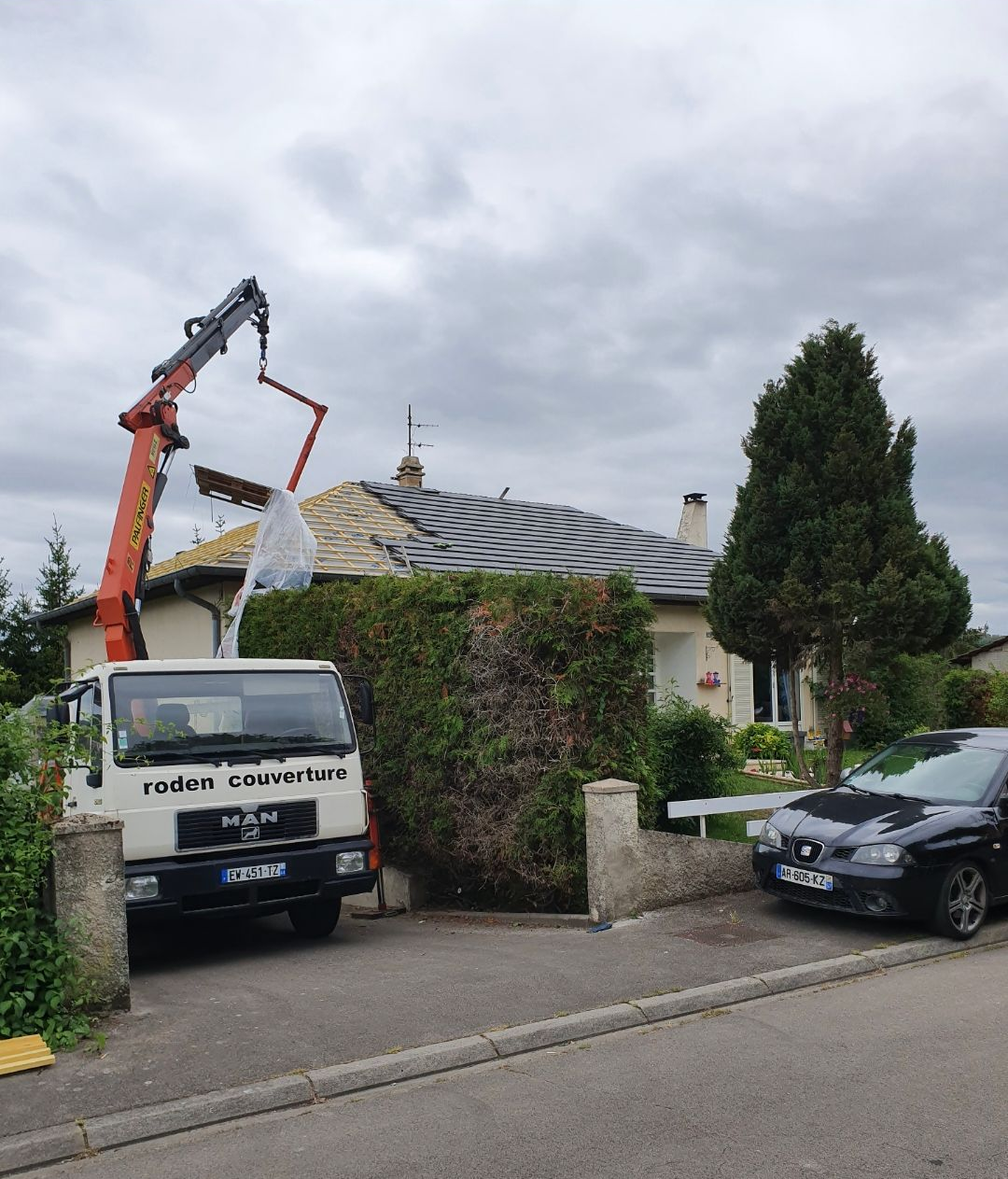 Remplacement de la toiture : grue au-dessus de la maison, camion dans l'allée, toiture partiellement remplacée, ciel couvert.