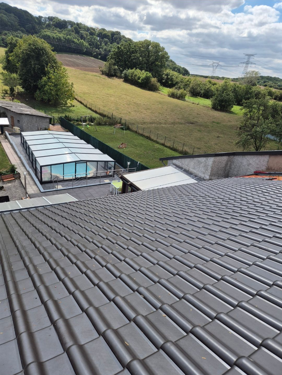 Vue d'un toit de tuiles sombres, donnant sur une piscine entourée d'une verrière et une colline herbeuse sous un ciel nuageux.