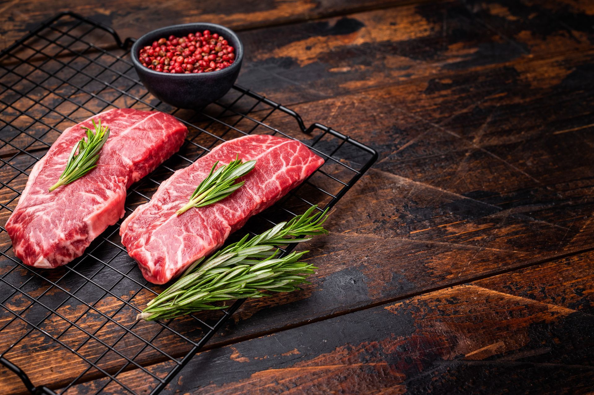 Deux steaks crus au romarin sur un gril, à côté d'un bol de grains de poivre sur une surface en bois.