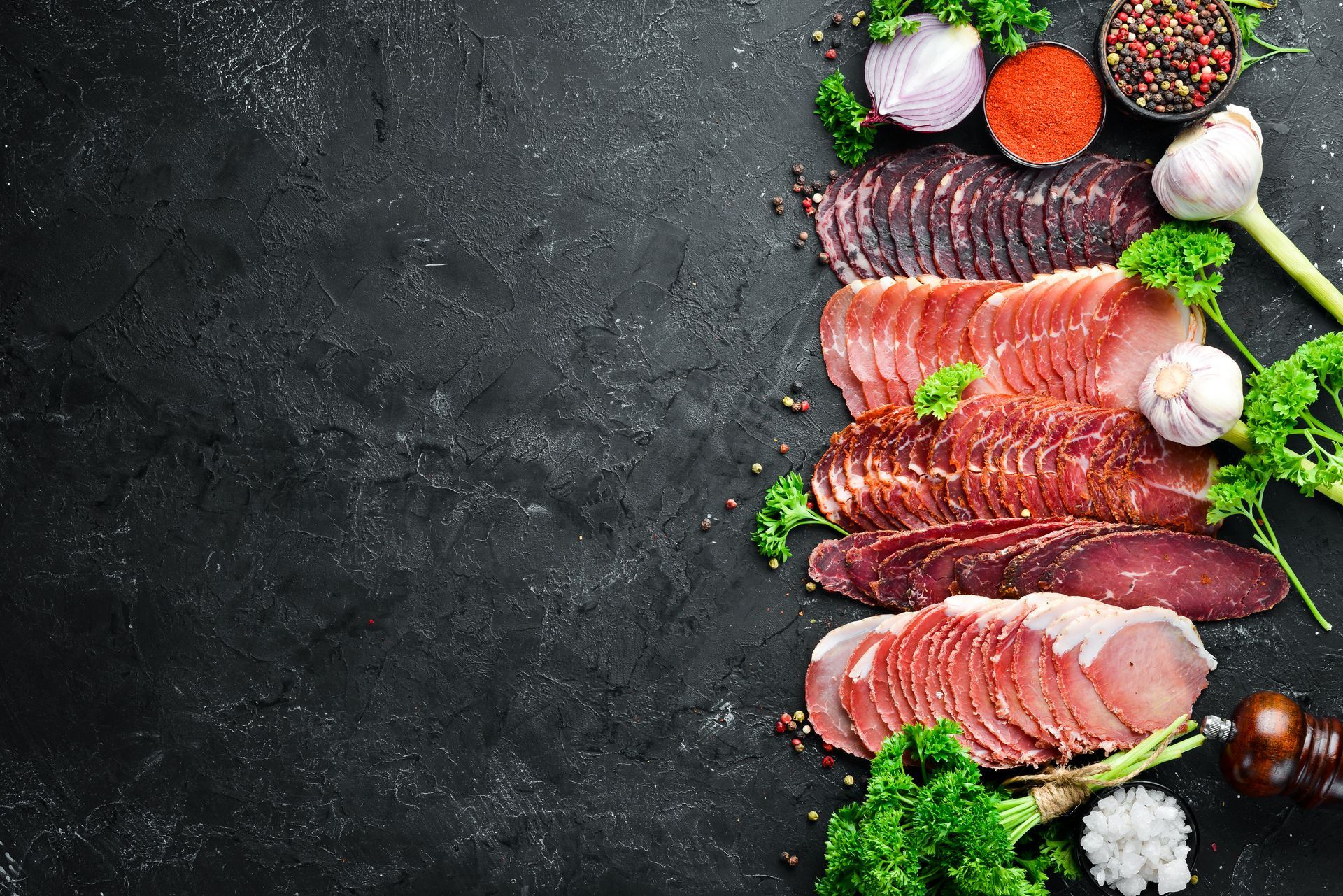 Assortiment de charcuteries tranchées avec de l'ail, des herbes et des épices sur une surface noire.