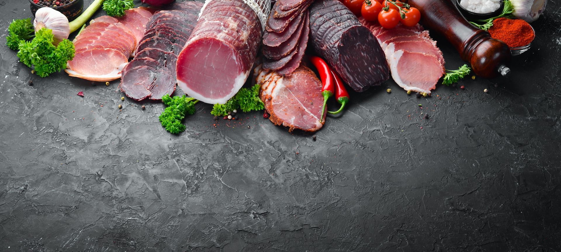 Assortiment de charcuteries disposées sur une surface en pierre foncée, accompagnées d'herbes fraîches, de tomates et d'épices.