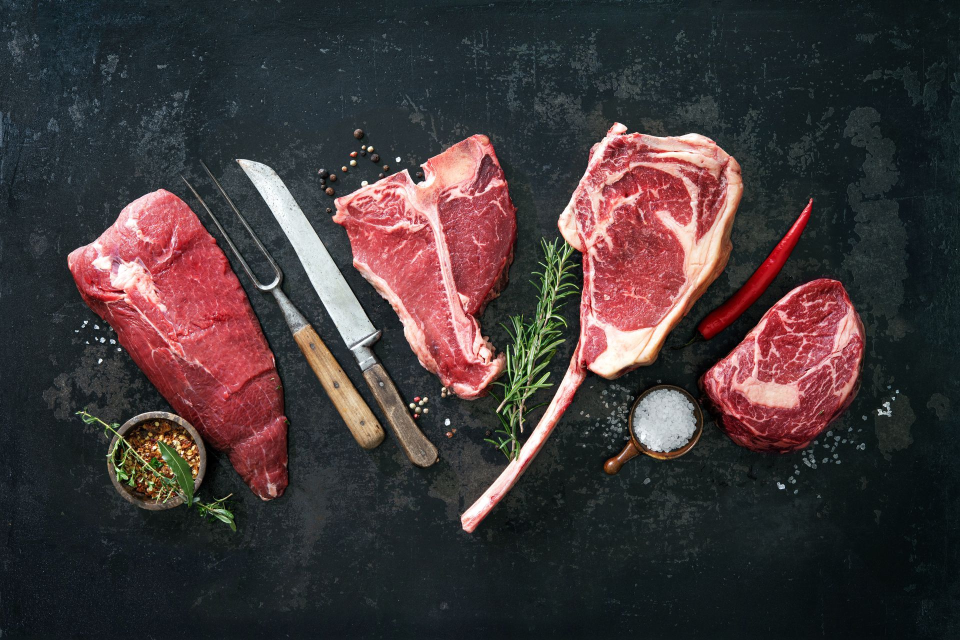Des morceaux de bœuf crus, y compris un steak en T, sont placés sur une surface sombre avec des assaisonnements, une fourchette à découper et un couteau.