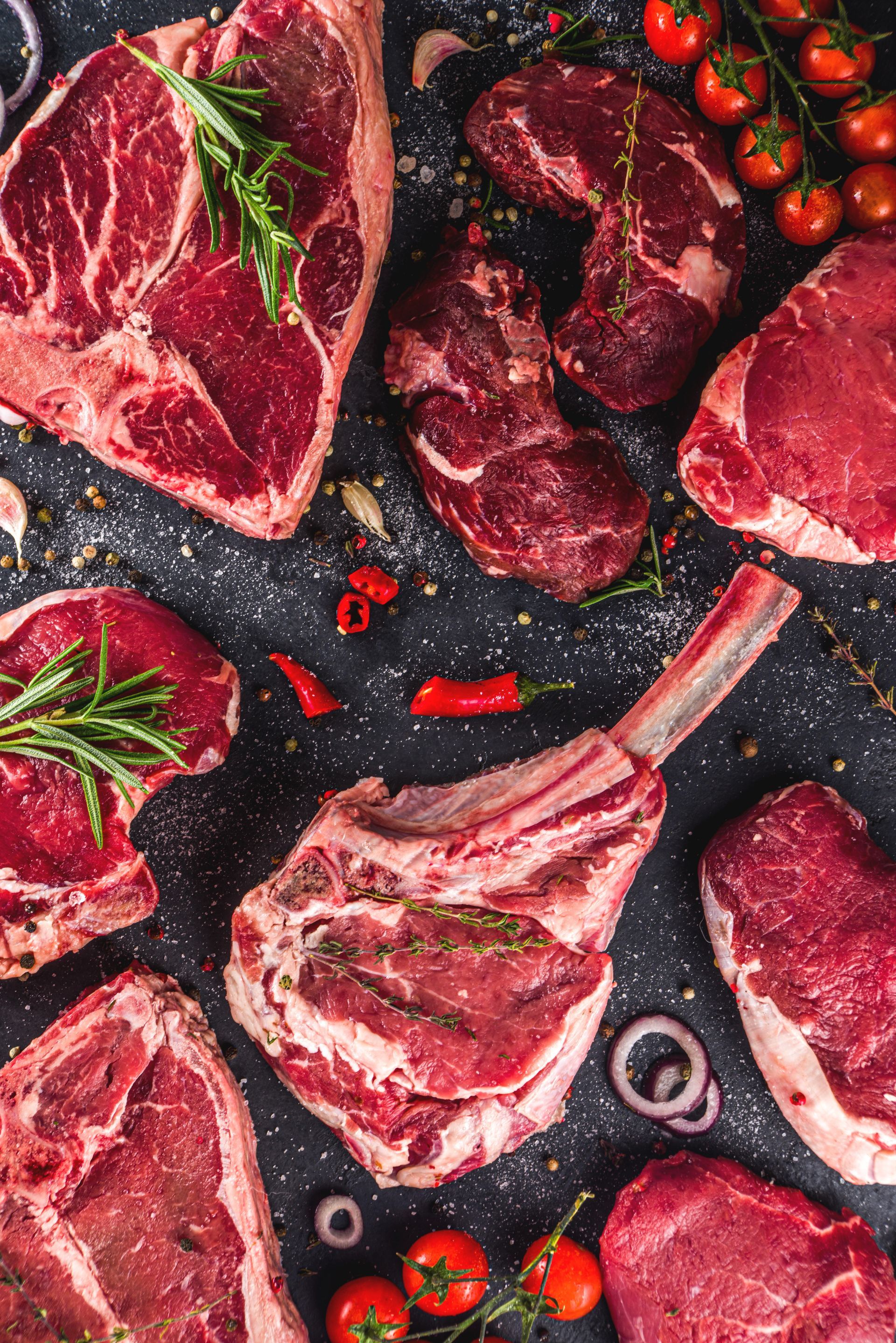 Divers morceaux de viande rouge crue, romarin, tomates cerises et piments sur une surface sombre.