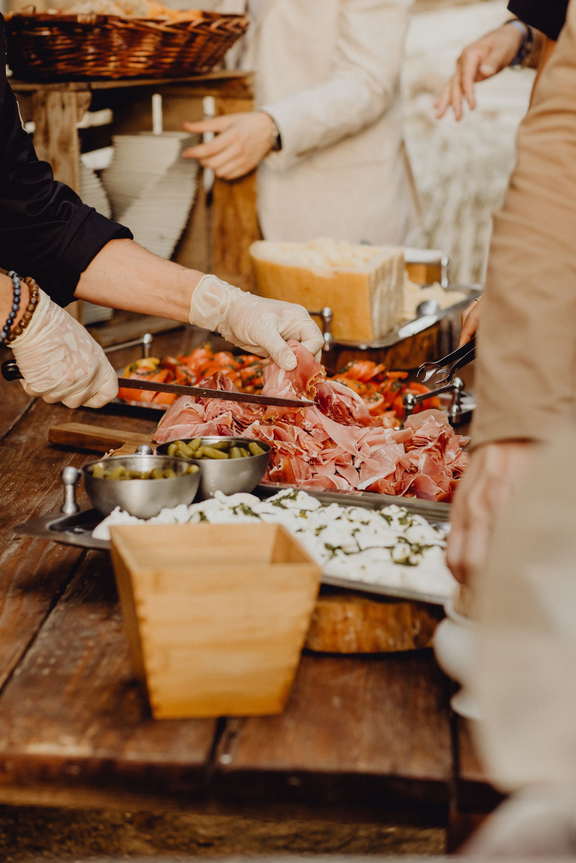 Buffet de nourriture : des mains gantées servent du prosciutto, des tomates et du fromage sur une table en bois rustique.