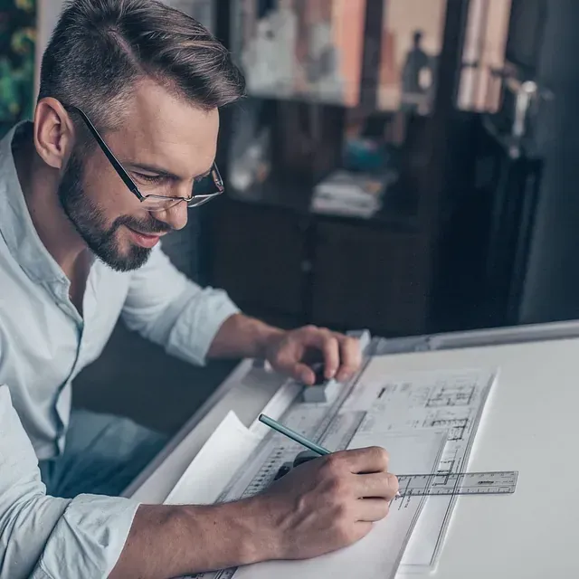 Un hombre con gafas, barba y camisa azul claro dibuja planos arquitectónicos en una mesa de dibujo. Sonríe.
