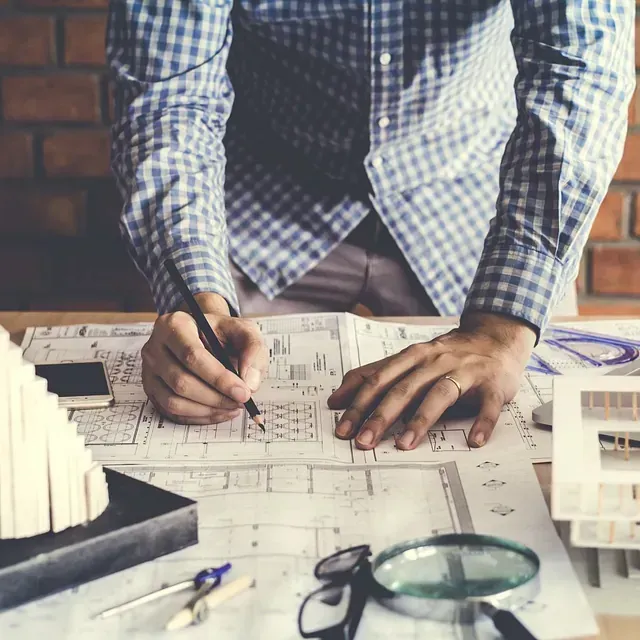 Arquitecto dibujando planos en un escritorio, rodeado de maquetas de edificios, herramientas y una lupa.