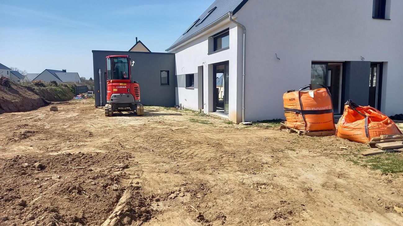 Chantier de construction avec une pelleteuse rouge à côté d'une maison blanche et des piles de matériaux de construction.