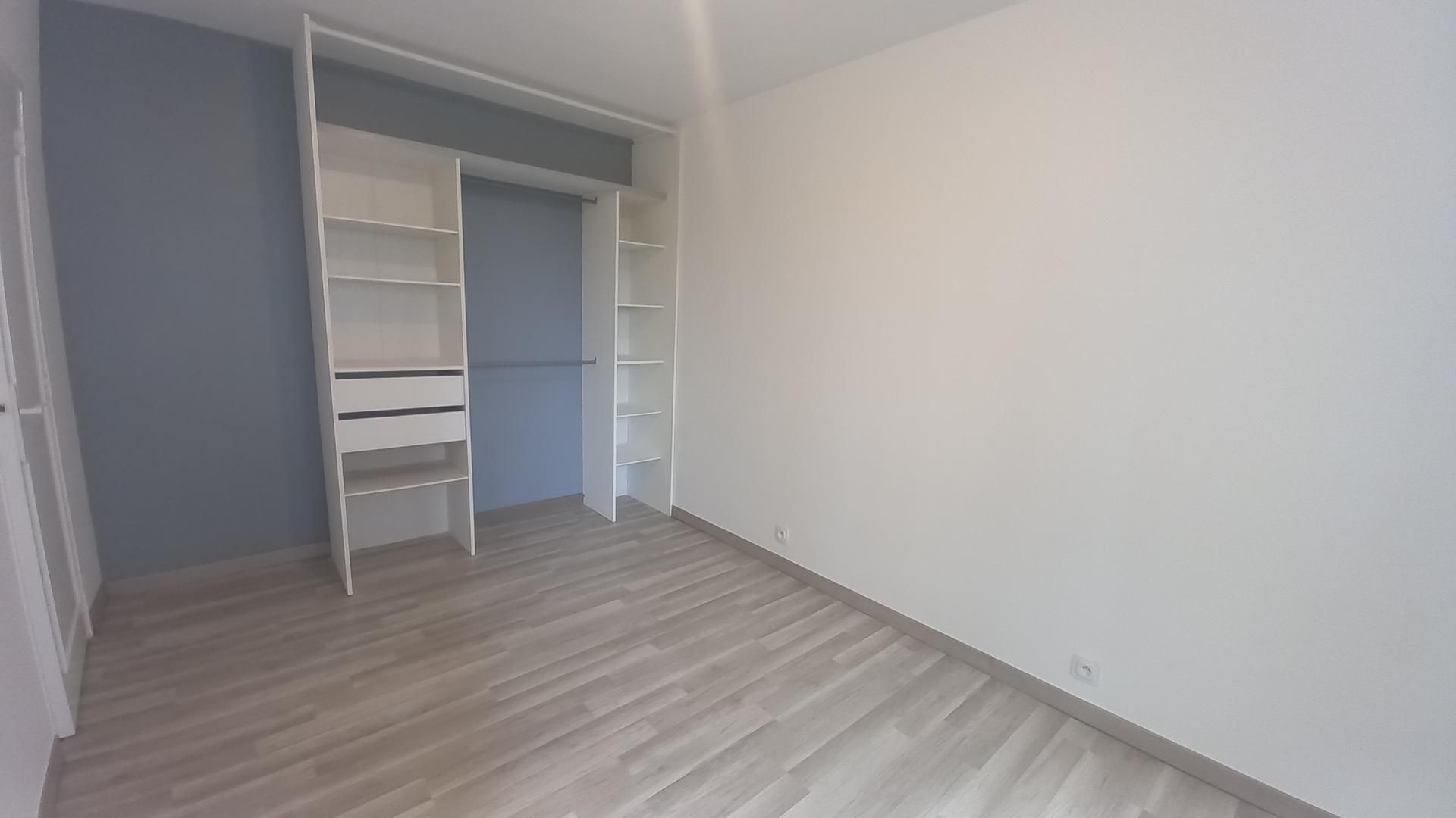 Chambre moderne vide avec parquet clair, murs blancs et étagères de placard ouvertes.