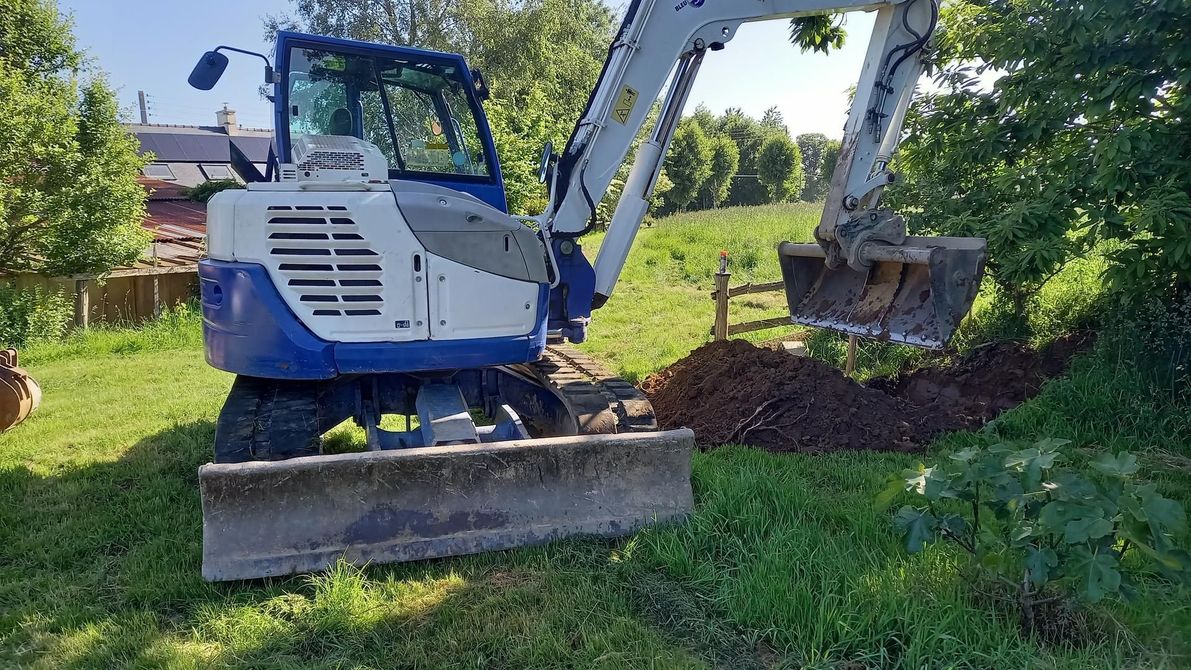 Une excavatrice à bras levé creuse la terre à côté d'une pelouse et d'arbres au soleil.
