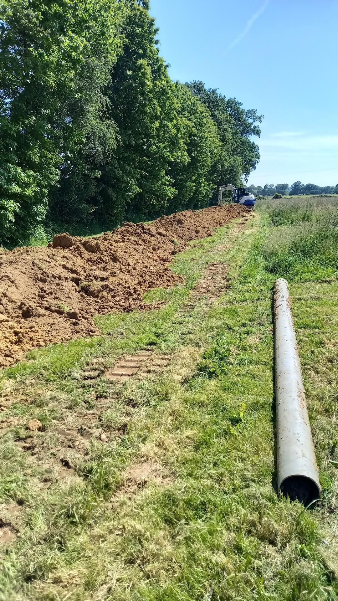 Un tas de terre borde un champ herbeux, près d'arbres, avec un gros tuyau gris au premier plan.