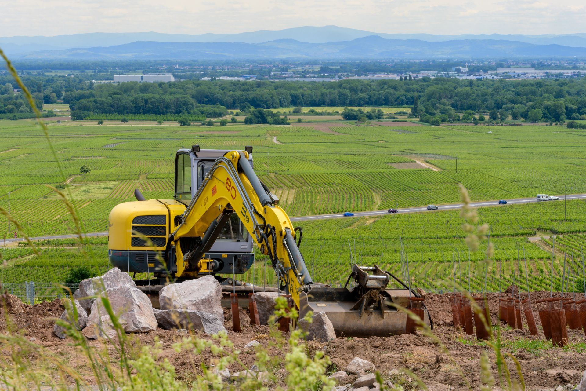 Une pelleteuse jaune creuse dans un champ herbeux avec des montagnes à l'horizon.