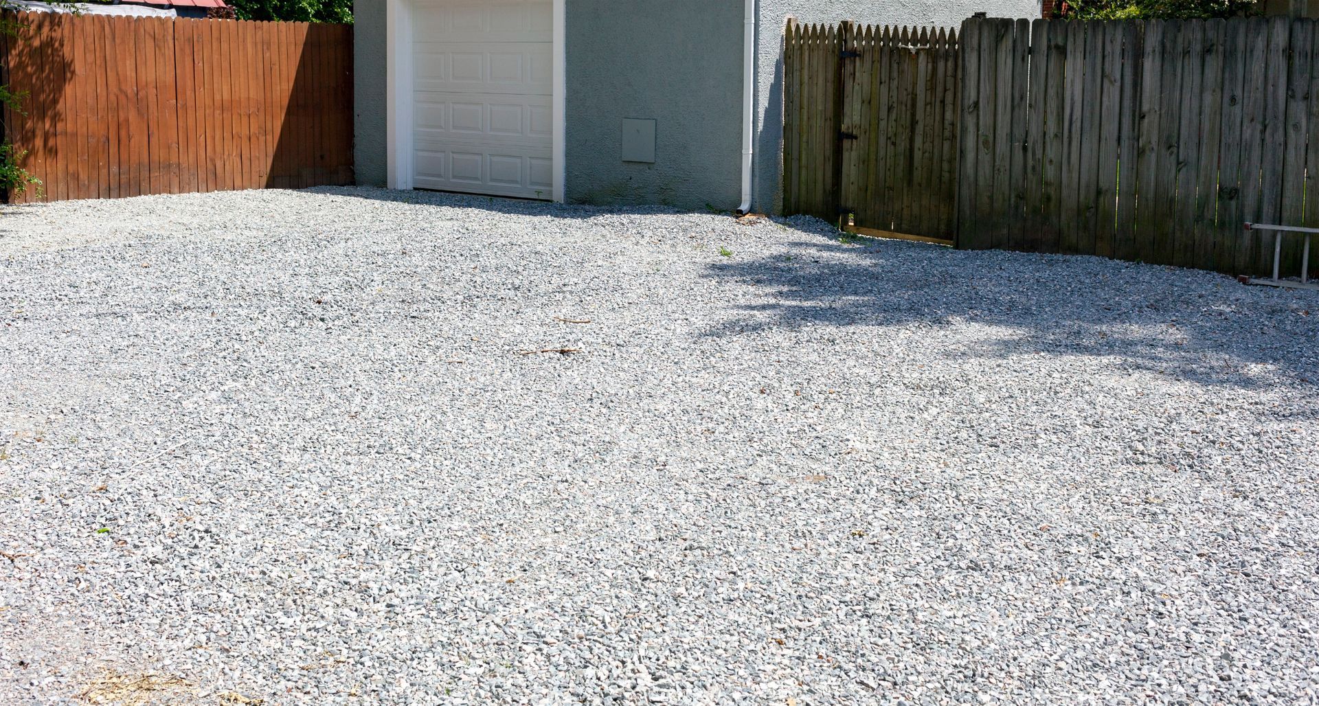 Allée de gravier longeant un garage blanc, bordée d'une clôture et d'un portail en bois.
