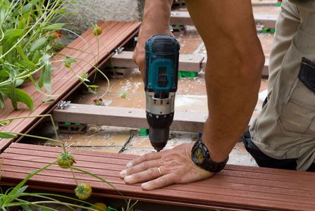 Une personne utilise une perceuse électrique pour fixer des planches sur une terrasse en bois extérieure.