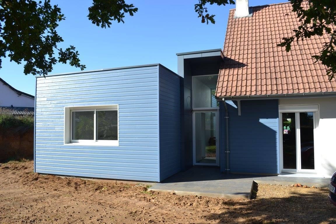Extension moderne bleue avec fenêtre, jouxtant une maison au toit de tuiles rouges.
