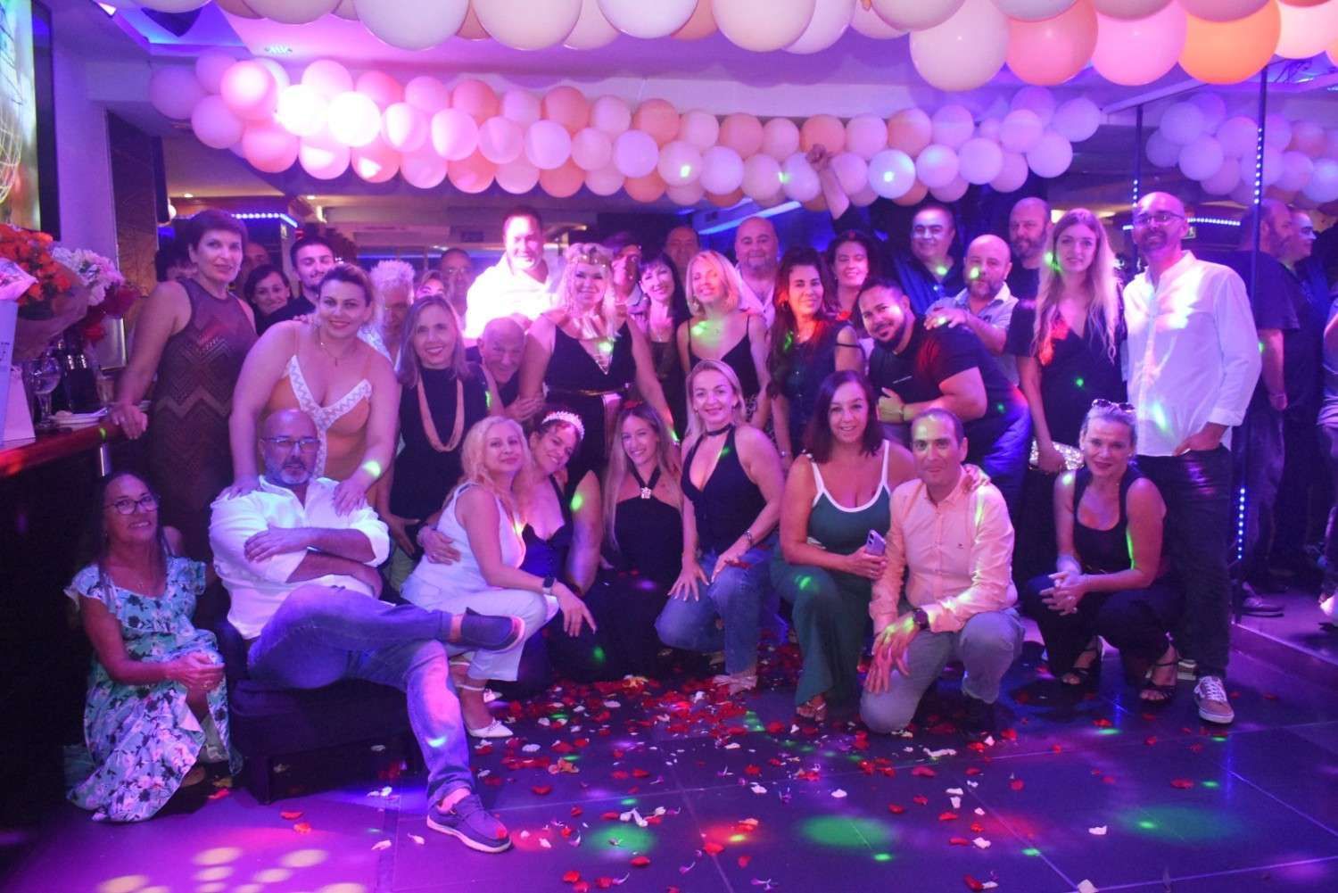 Un numeroso grupo de personas posando para una foto en una fiesta en interiores decorada con globos rosas y blancos e iluminación de escenario.