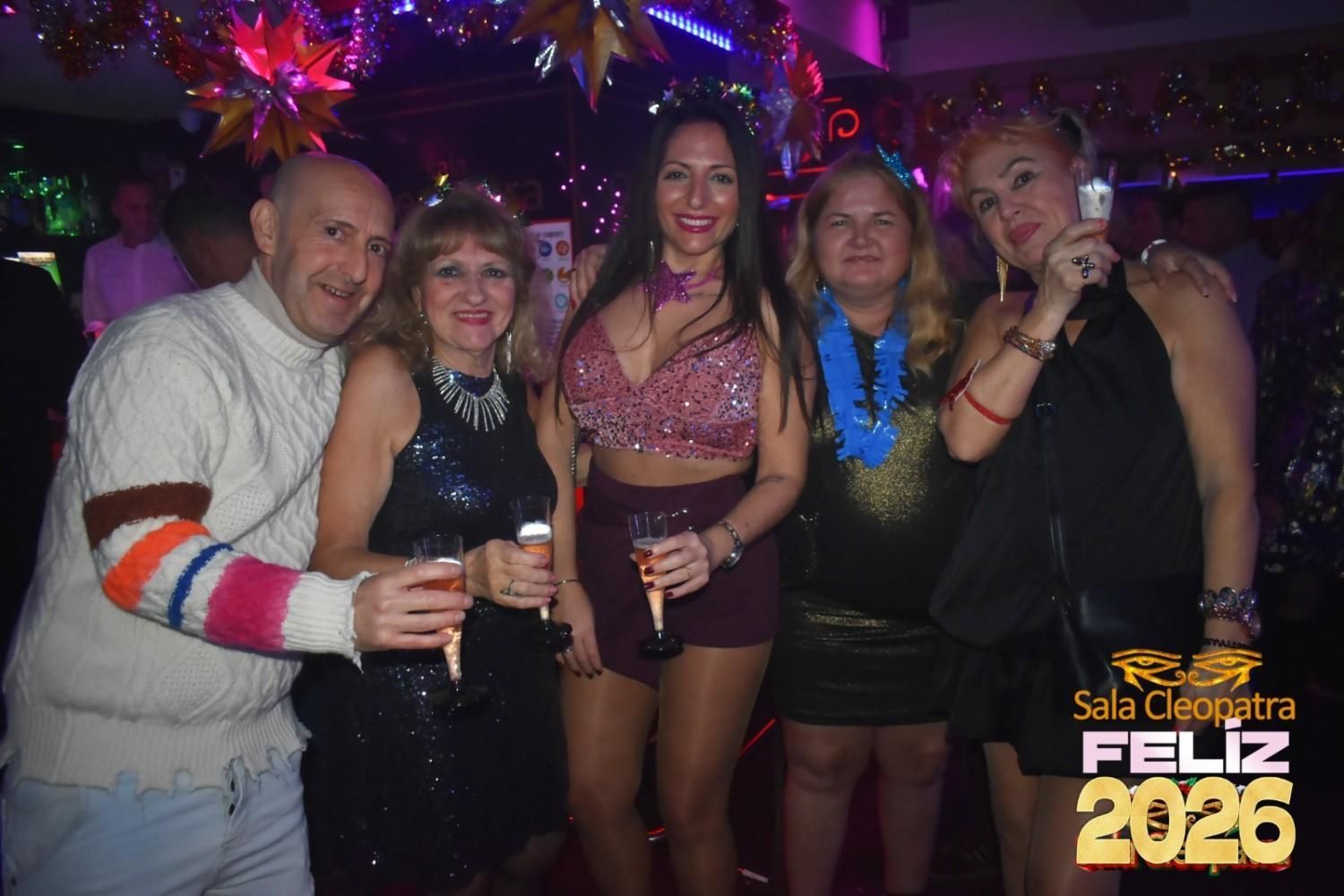 Un grupo de cinco personas posa para una foto festiva en una discoteca, sosteniendo bebidas con un gráfico que dice 