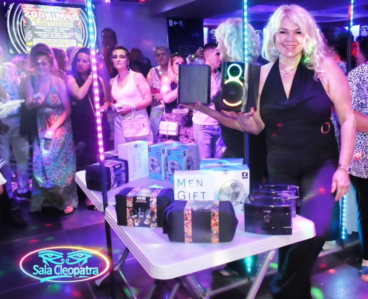 En una fiesta, una persona sonriente se encuentra detrás de una mesa, sosteniendo una caja de altavoces junto a otros artículos de regalo para un sorteo.