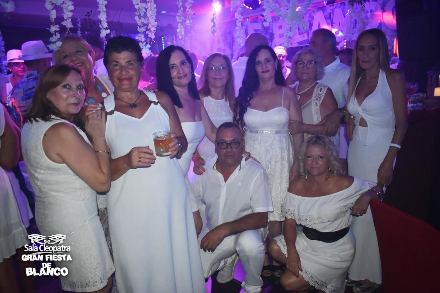 Un grupo de personas vestidas de blanco en un evento nocturno en interiores con iluminación morada y decoración festiva.