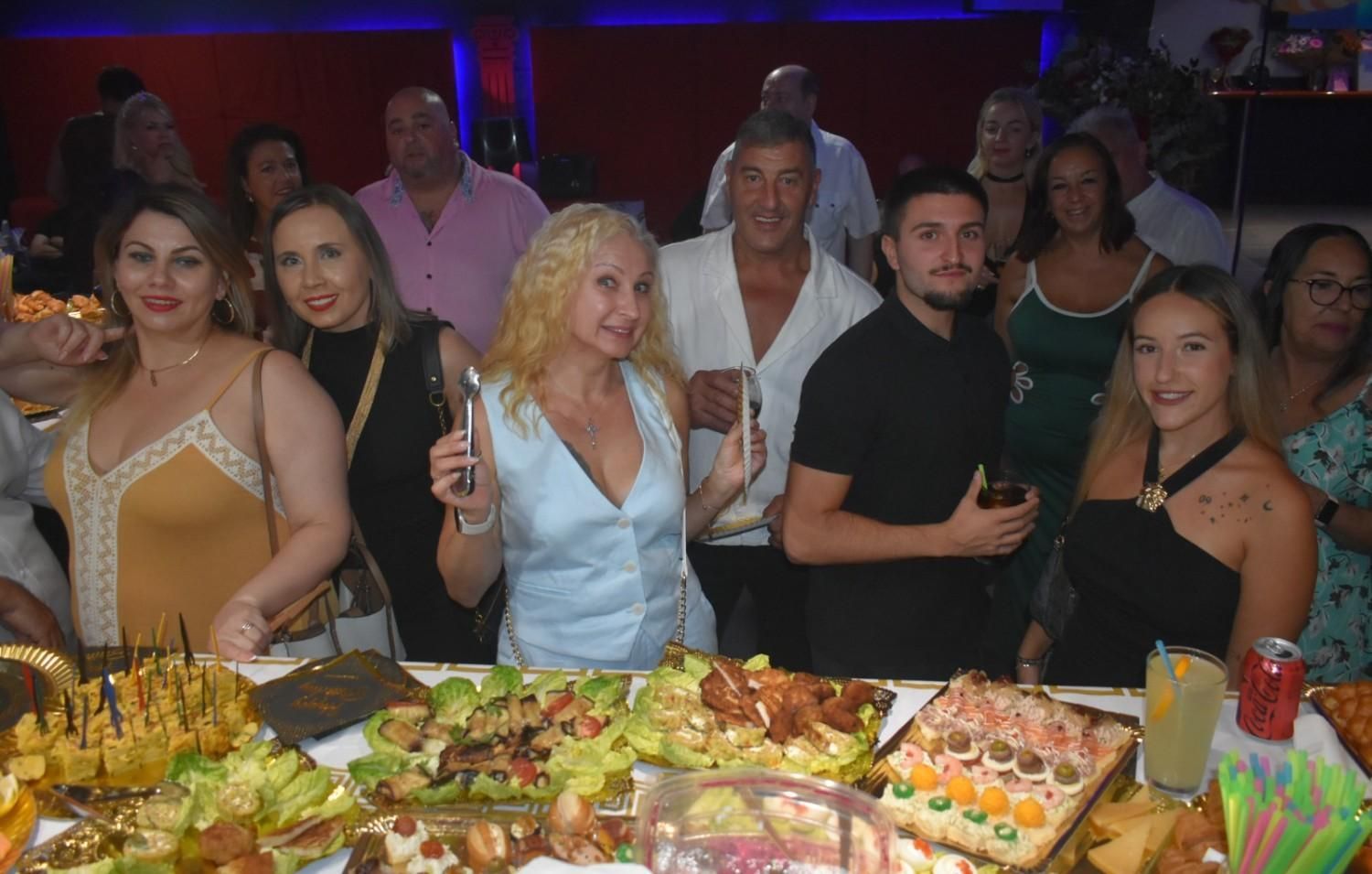 Un grupo de personas de pie detrás de una mesa repleta de aperitivos y bocadillos variados en un evento nocturno en un espacio cerrado.