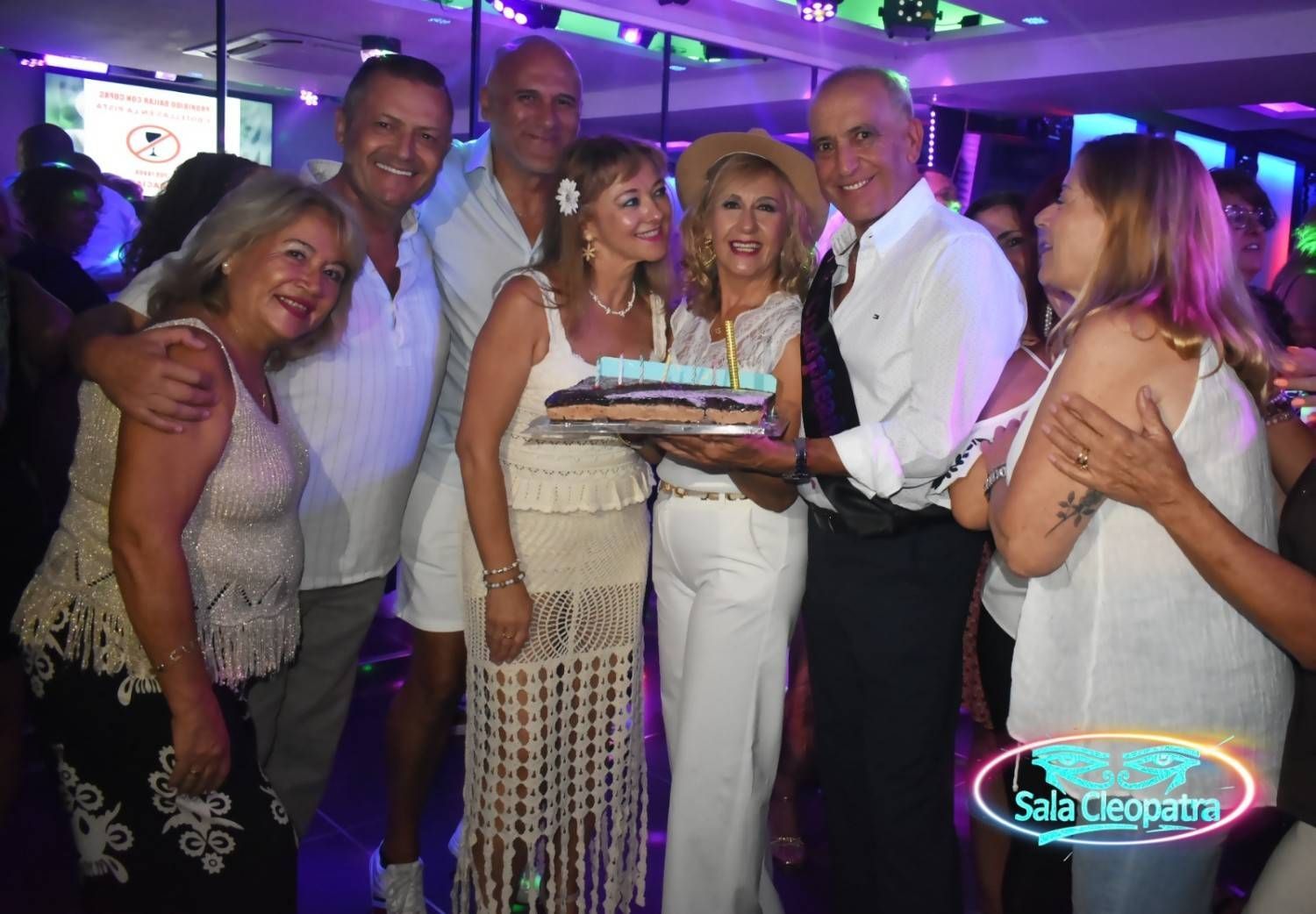 Un grupo de personas sonríe mientras se reúnen alrededor de una tarta de cumpleaños en un local con iluminación tenue y de tonos morados.