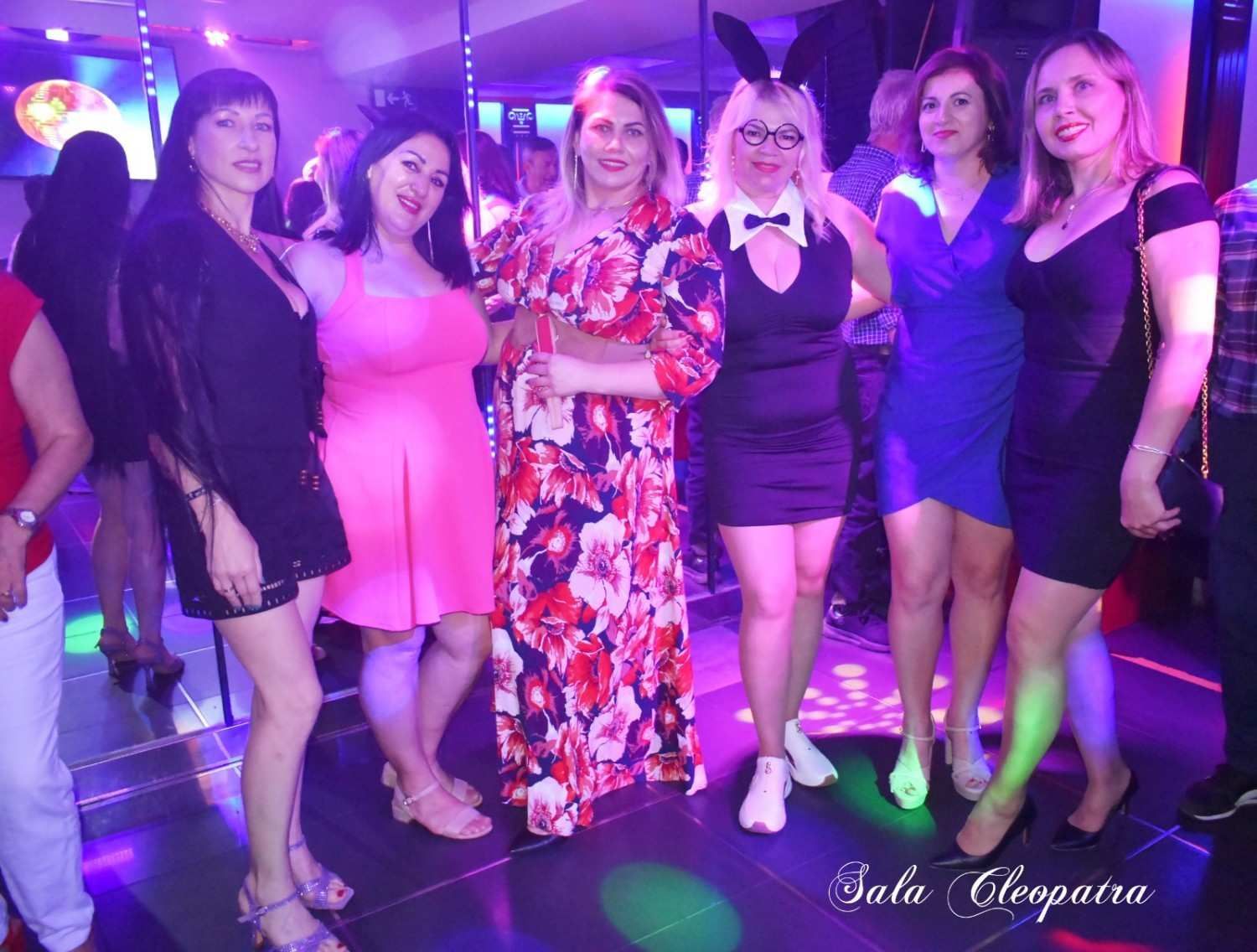 Seis personas haciendo fila en un club con poca luz y luces de colores en el suelo, posando para una foto.