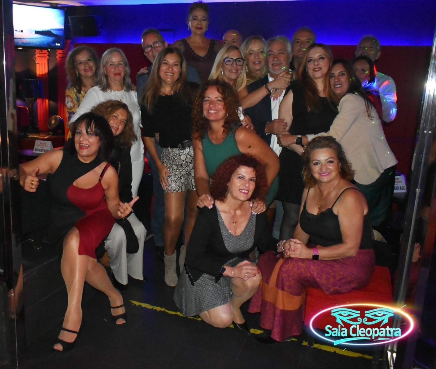 Un grupo numeroso y sonriente posa para una foto en un local con poca luz, con el logotipo de la Sala Cleopatra en la parte inferior.