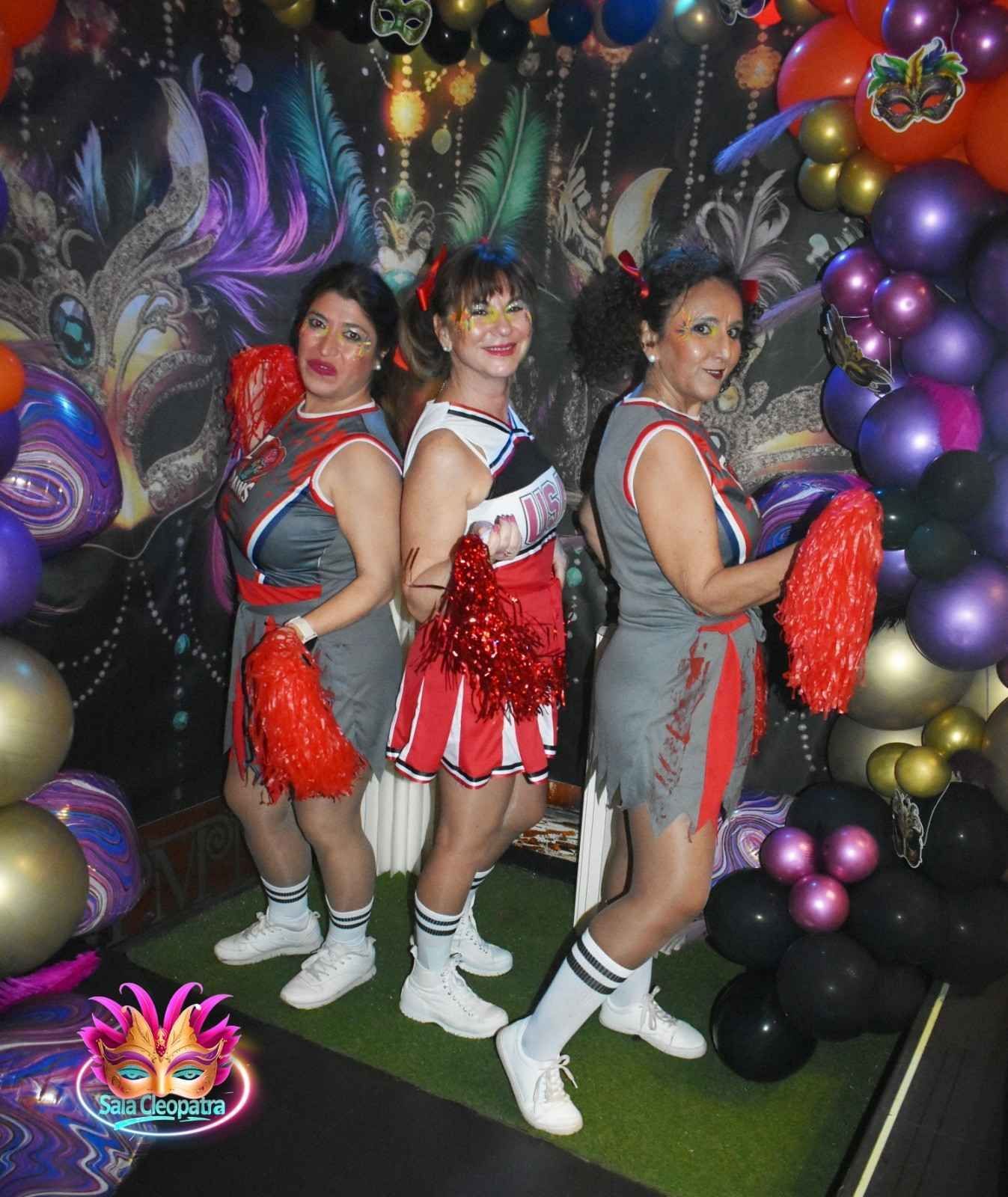 Tres personas vestidas con uniformes de animadoras grises y rojos, con pompones rojos, posan frente a un fondo festivo con globos.