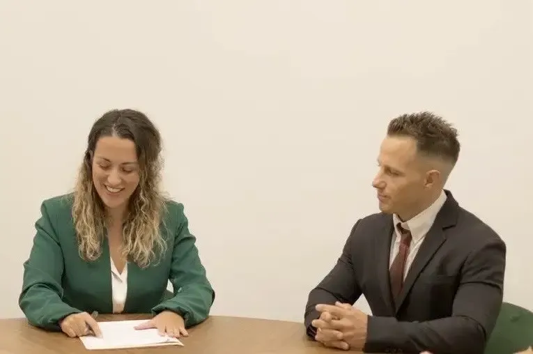 Un profesional escribe mientras consulta con una pareja en un escritorio, en un entorno de oficina con papel tapiz tropical.