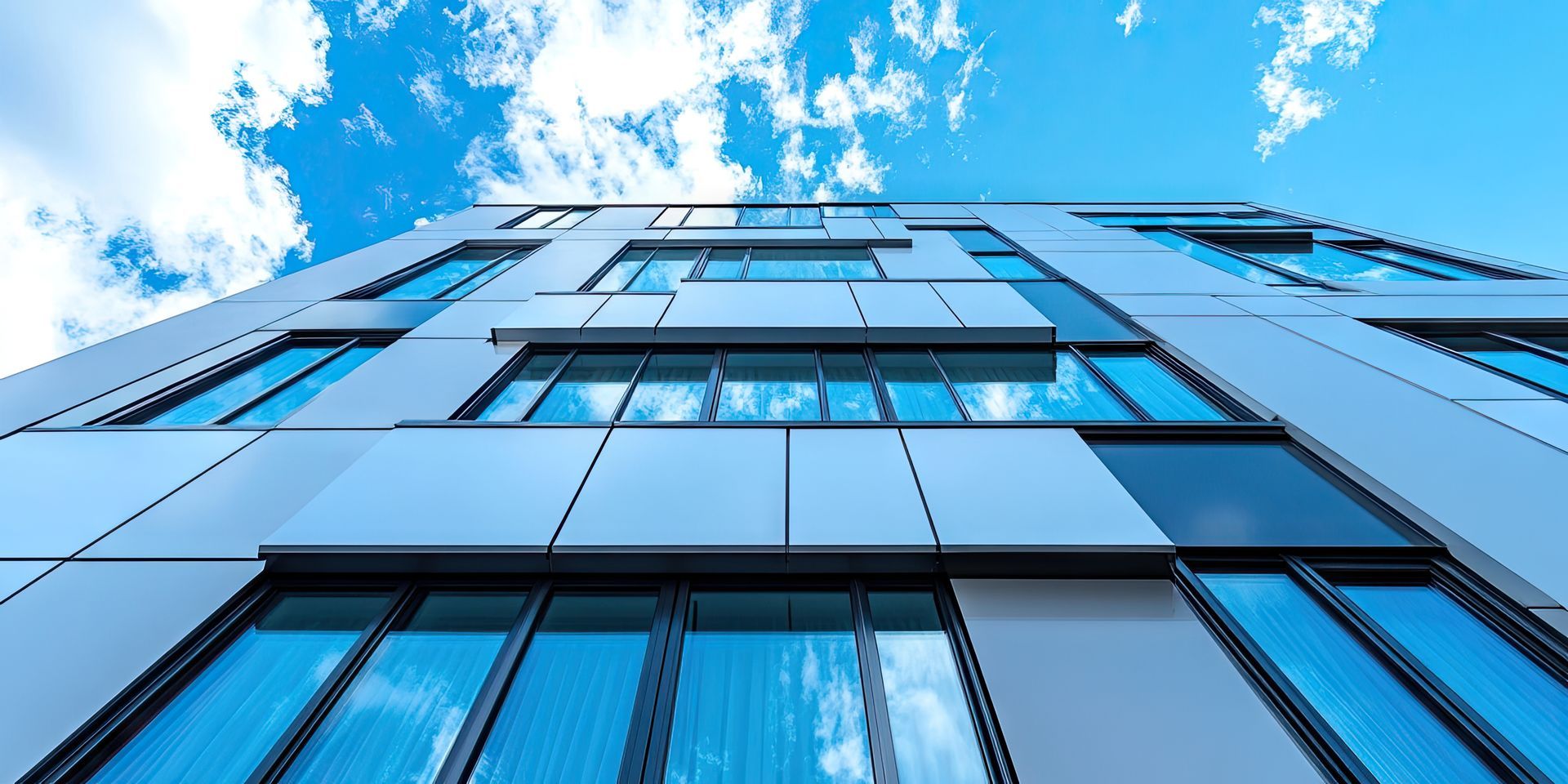Vue en contre-plongée d'un bâtiment moderne aux fenêtres bleues et aux panneaux blancs, se détachant sur un ciel bleu clair nuageux.