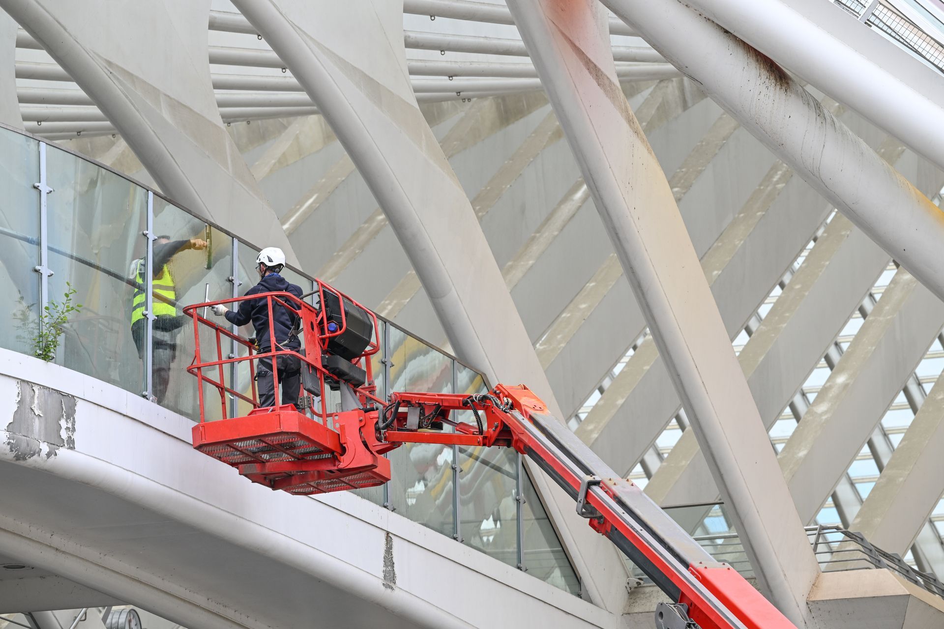 Des ouvriers nettoient des panneaux de verre dans une nacelle sur un immeuble moderne blanc à poutres métalliques.