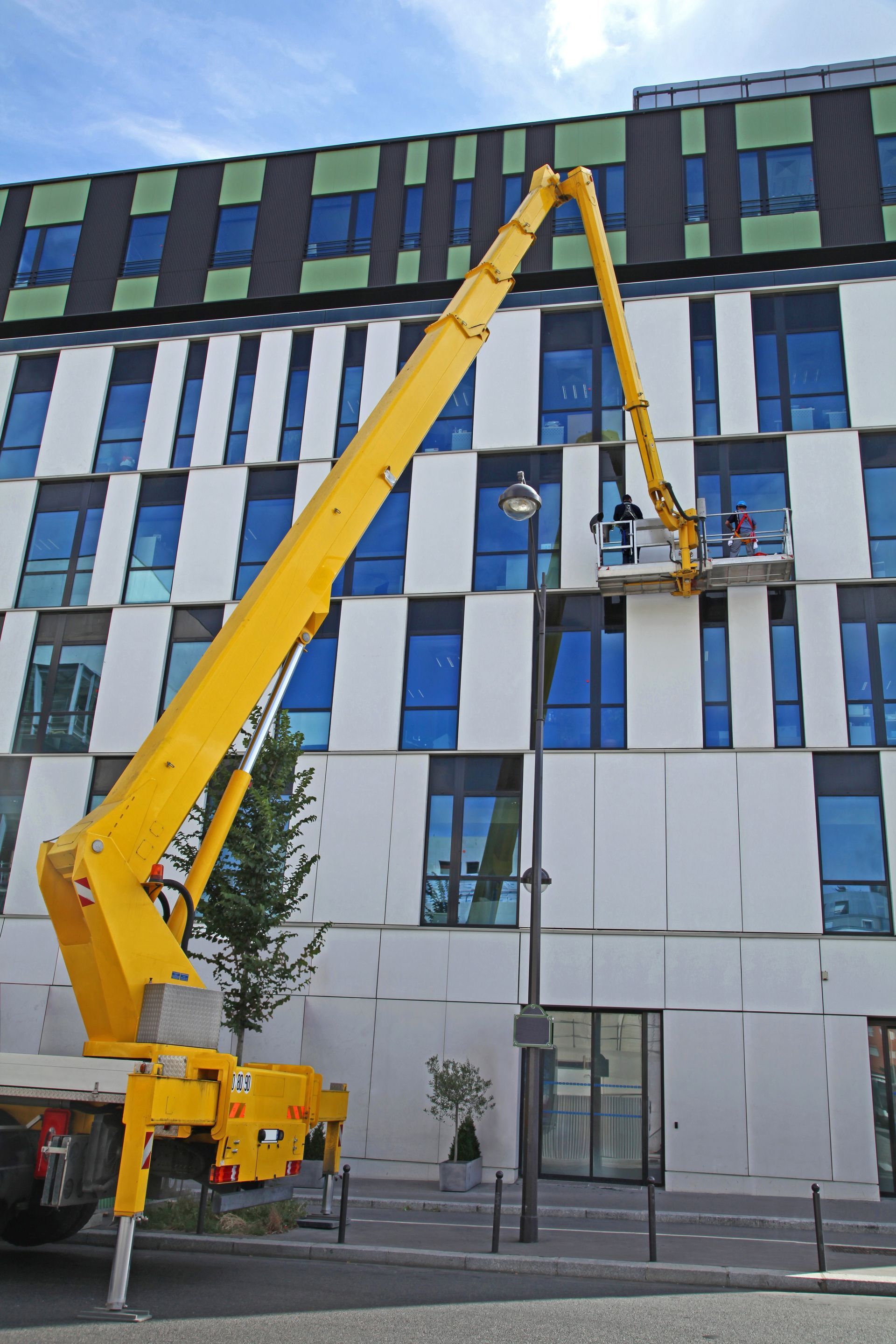 Une nacelle élévatrice jaune se déploie vers un bâtiment ; deux personnes se trouvent dans la nacelle. Façade bleue et blanche.