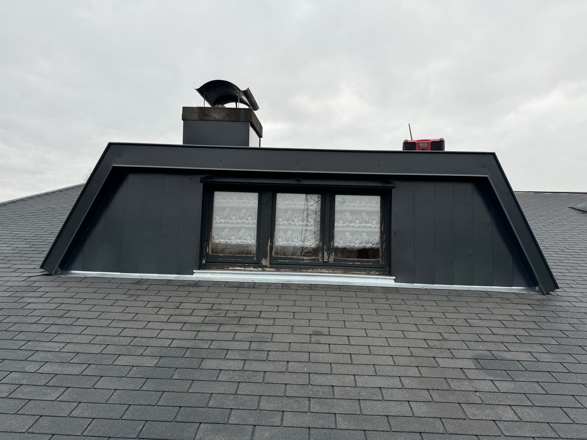 Lucarne gris foncé avec cheminée sur un toit de bardeaux sombres, se détachant sur un ciel nuageux.