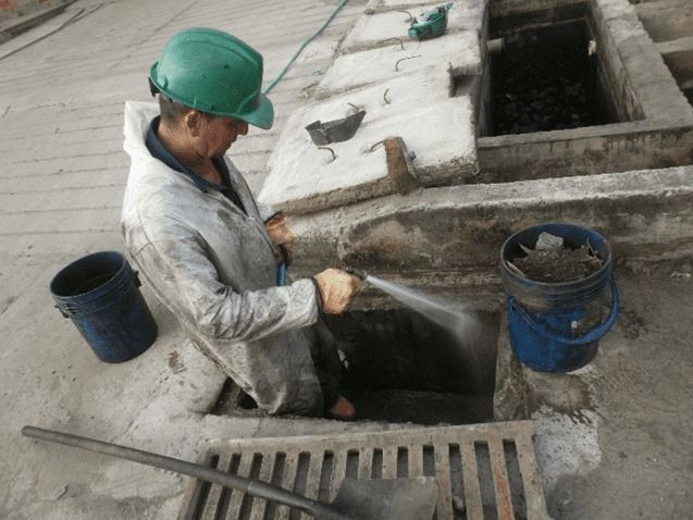Un hombre que lleva un casco verde está limpiando un desagüe con una manguera.