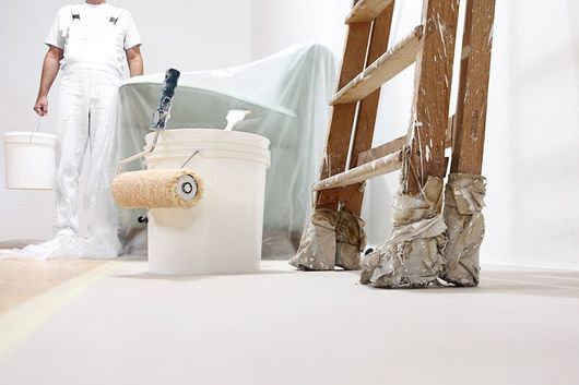 Pintor con mono blanco, cubo de pintura y rodillo, cerca de una escalera y muebles cubiertos, en una habitación que se está pintando.