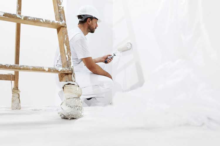 Pintor con overol blanco y casco pintando una pared con rodillo. Cerca hay una escalera de madera.