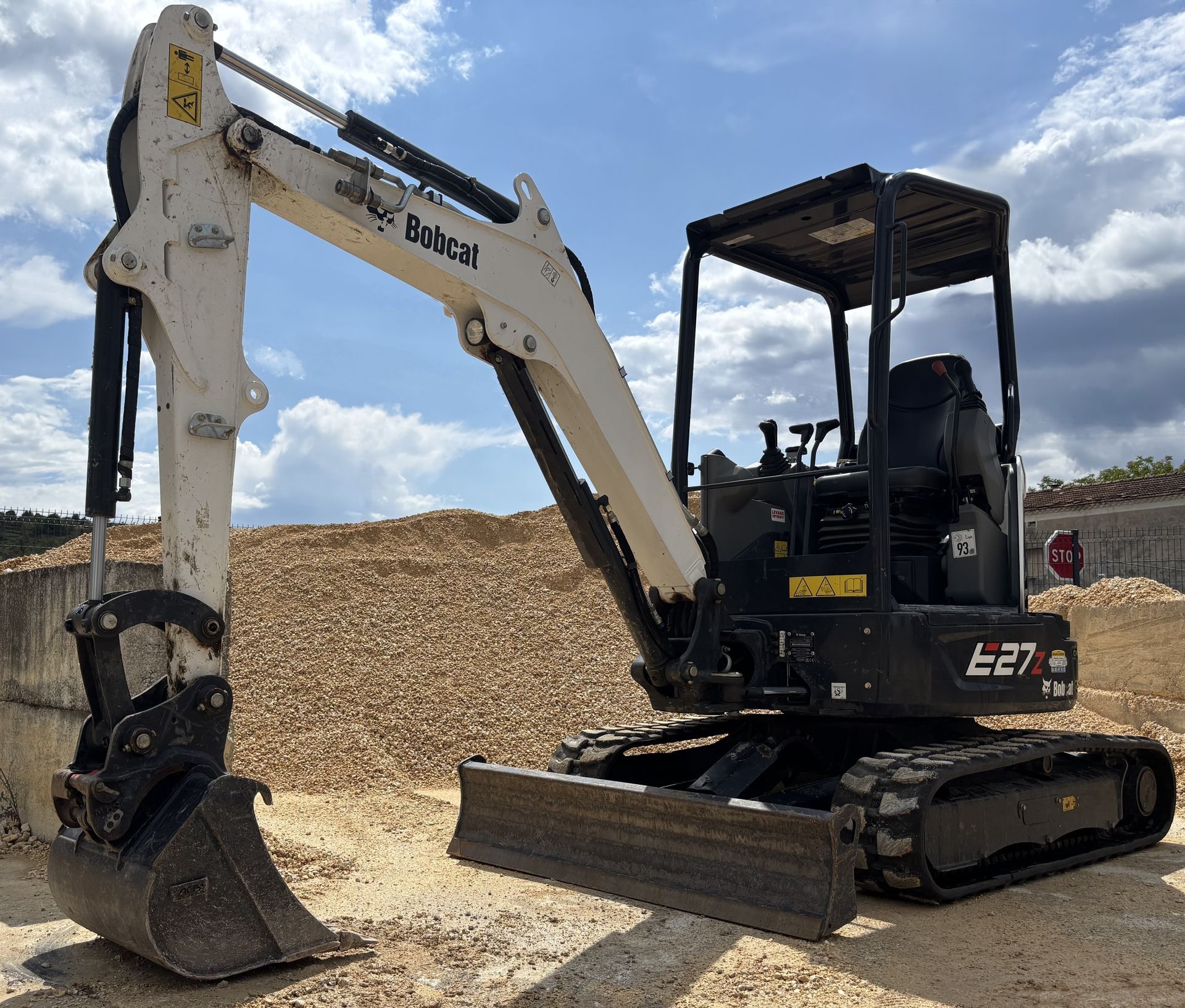 Excavatrice blanche et noire sur un terrain entouré de tas de matériaux avec un habitacle ouvert
