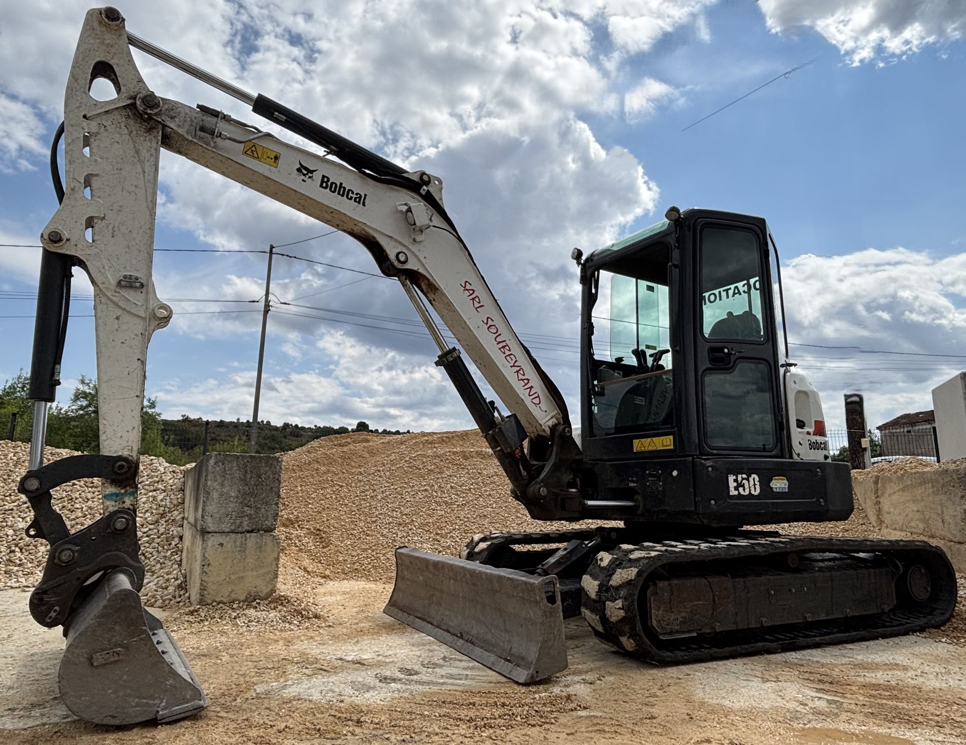 Excavatrice blanche et noire sur un terrain entouré de tas de matériaux avec un habitacle fermé