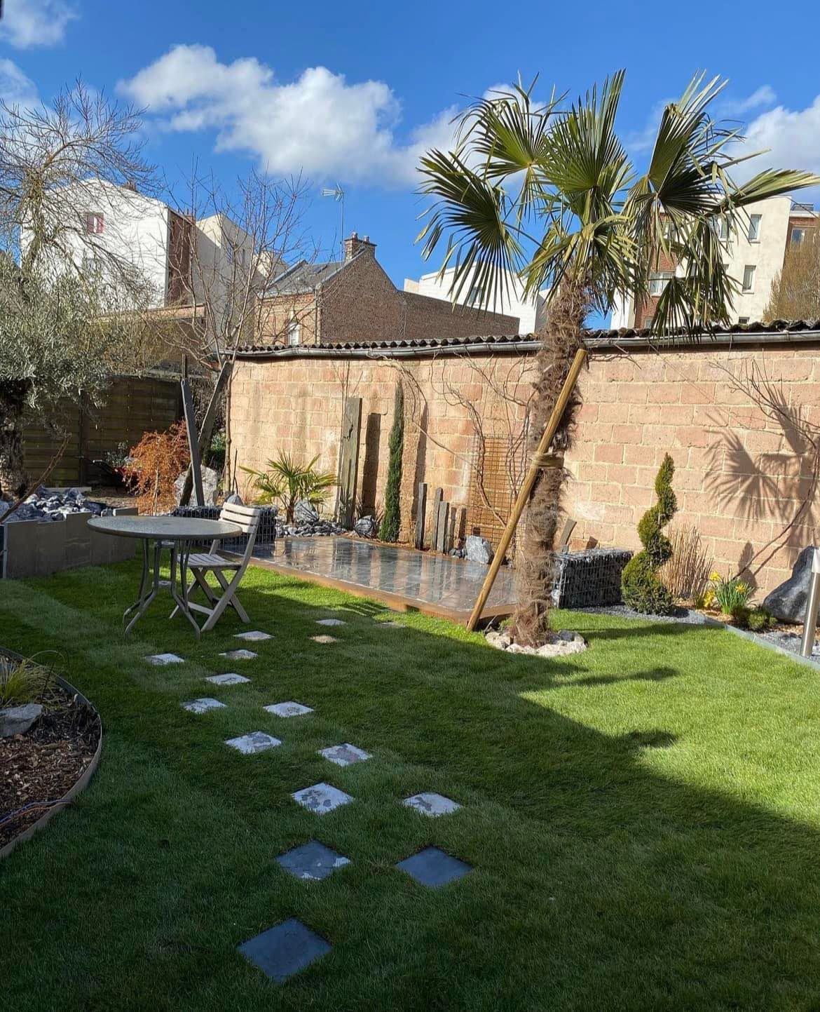 Un jardin ensoleillé avec une pelouse verdoyante, un chemin de pierre, un palmier et une petite table à manger adossée à un mur de briques.