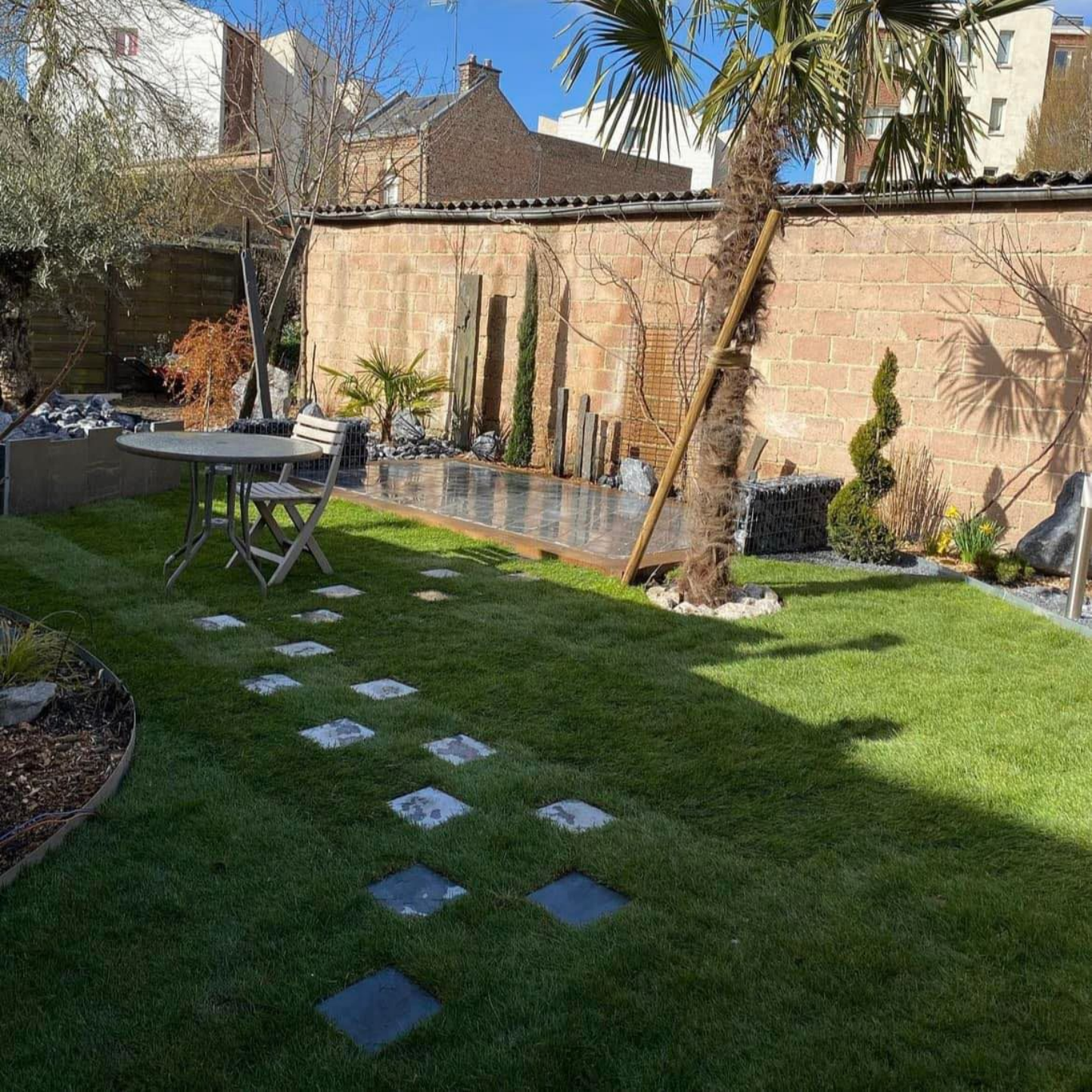 Un jardin ensoleillé avec une pelouse verte, des pas japonais, un petit ensemble table et chaises de bistrot et un mur de briques orné de palmiers.