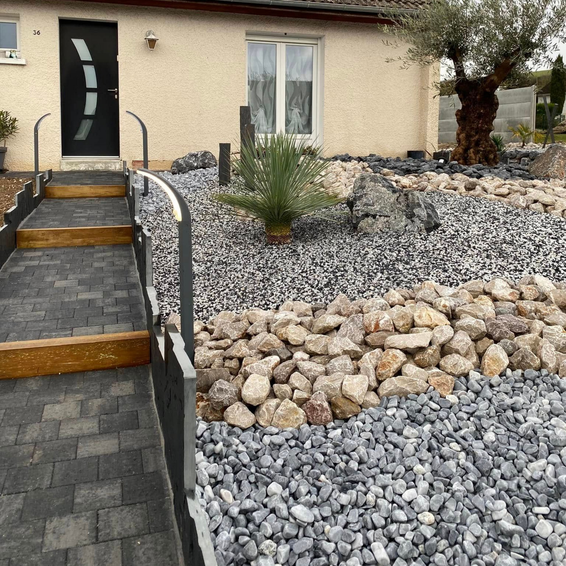 Une entrée de maison moderne avec des escaliers pavés de pierre, une rampe en métal et un jardin paysager avec du gravier gris et beige.