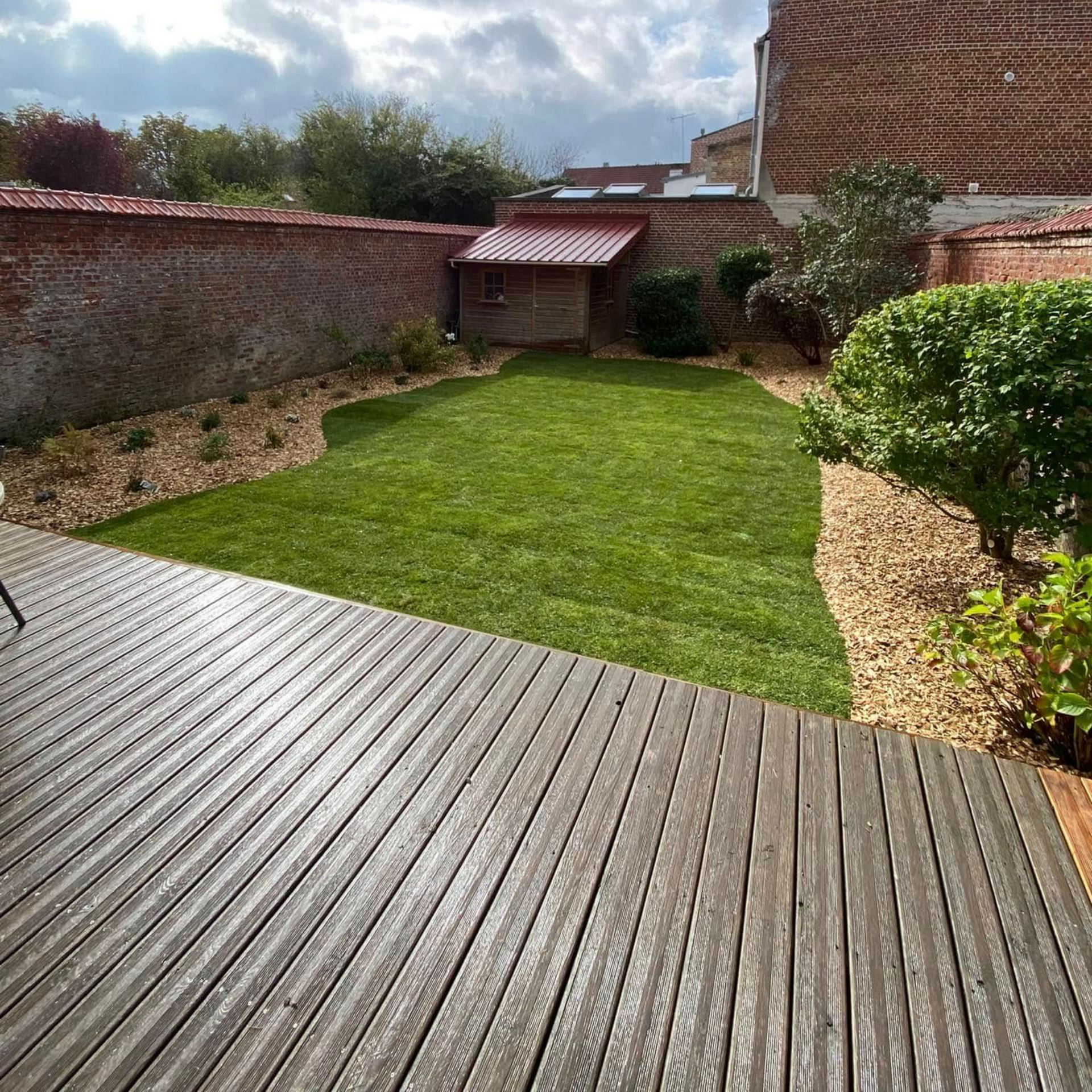 Une terrasse en bois surplombe un jardin gazonné bordé de gravier, avec un petit abri de jardin et un mur de briques rouges, sous un ciel nuageux.