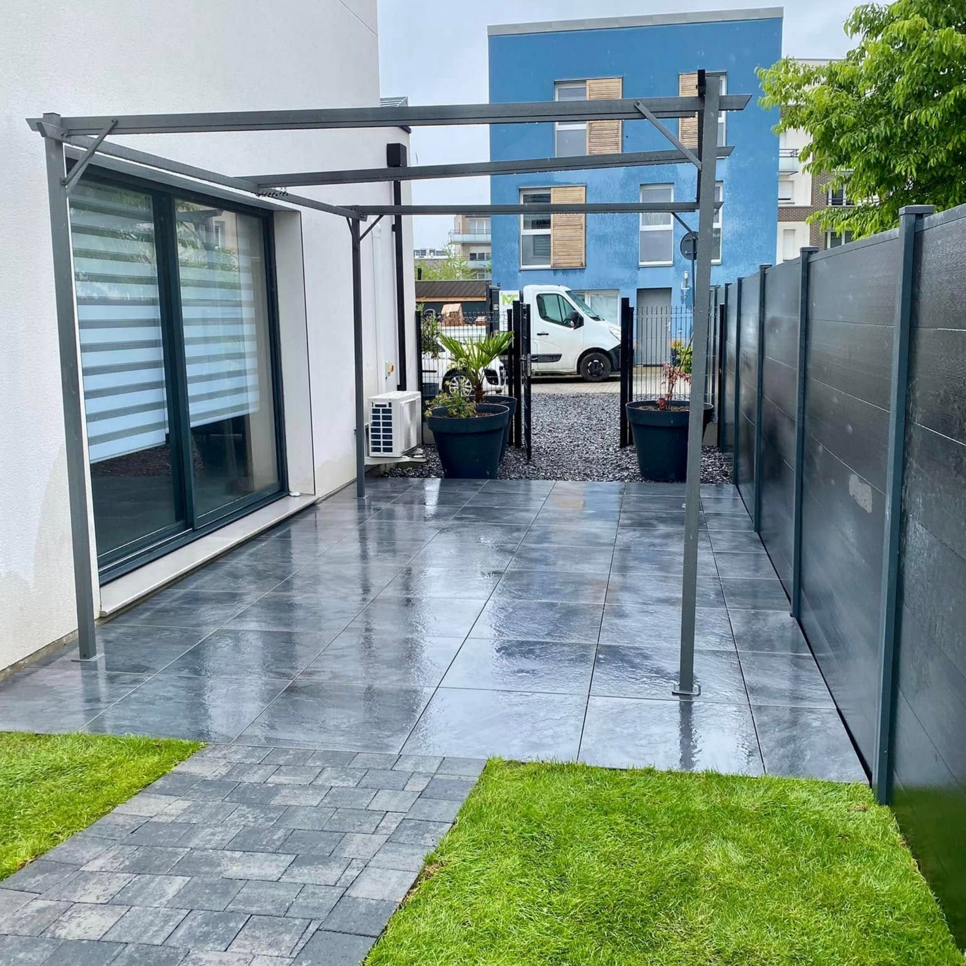 Une terrasse avec des dalles d'ardoise grise, une pergola en métal et une haute clôture pour préserver l'intimité, jouxtant une maison dotée d'une grande fenêtre.
