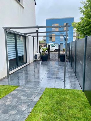 Un patio avec une pergola métallique recouvrant des dalles de pierre sombres, situé à côté d'un mur de maison blanc et d'une haute clôture sombre.