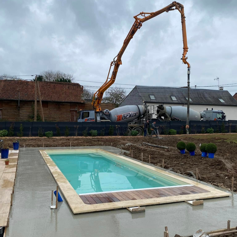 Un camion-pompe à béton déverse du ciment autour d'une piscine rectangulaire nouvellement installée sur un chantier de construction dans une cour arrière rurale.