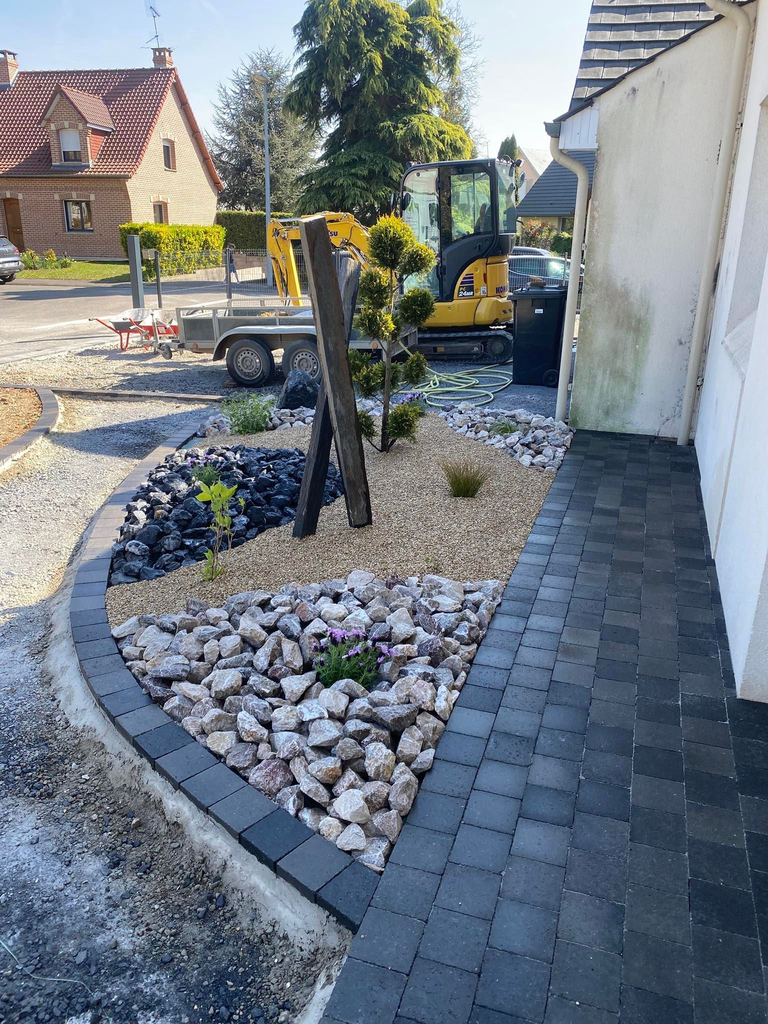 Un parterre de jardin paysager moderne avec des roches colorées et une allée pavée sombre, avec une pelleteuse de chantier à proximité.