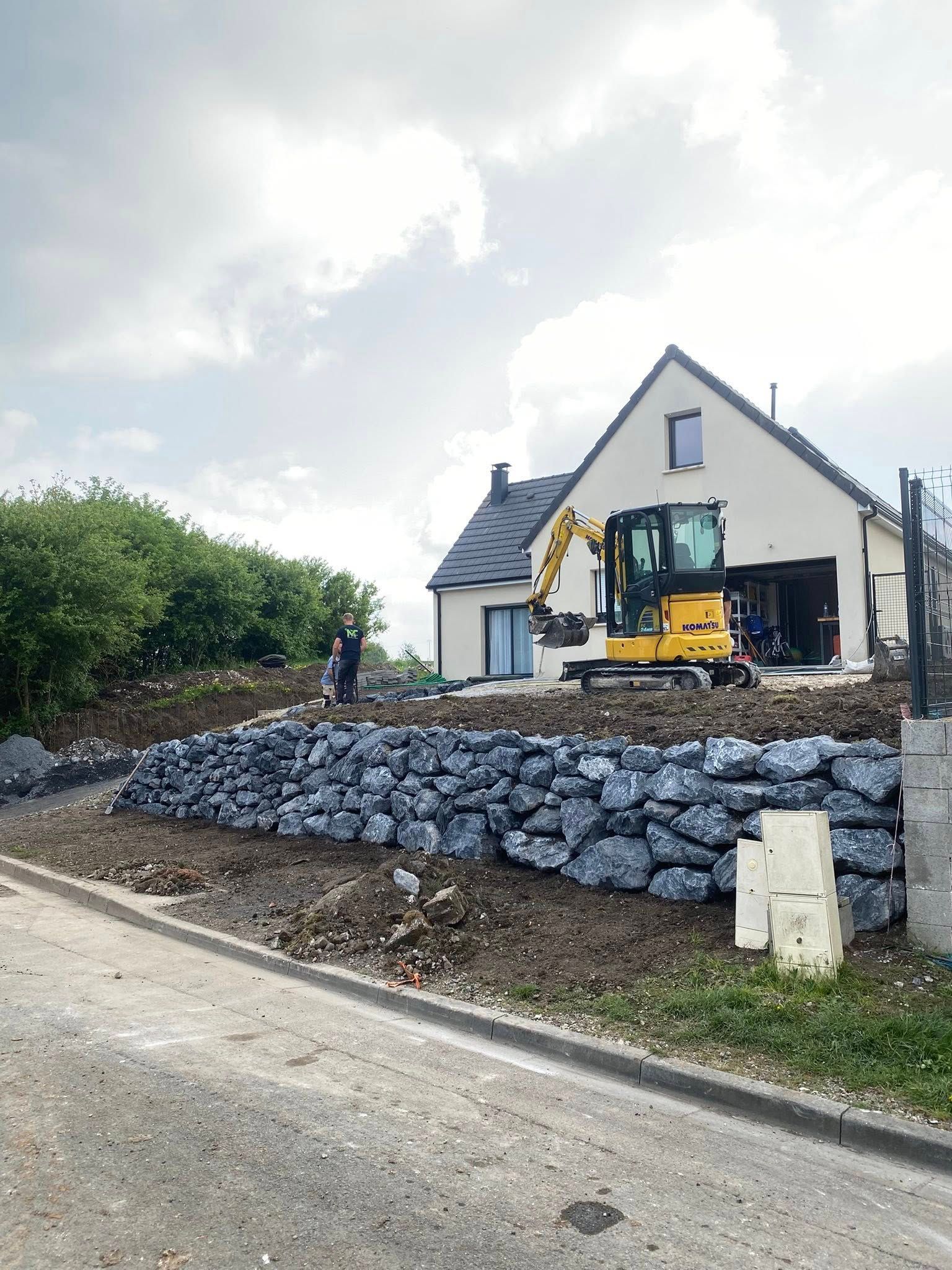 Une pelleteuse jaune travaille sur un mur de soutènement en pierre paysagé devant une maison, sous un ciel nuageux.