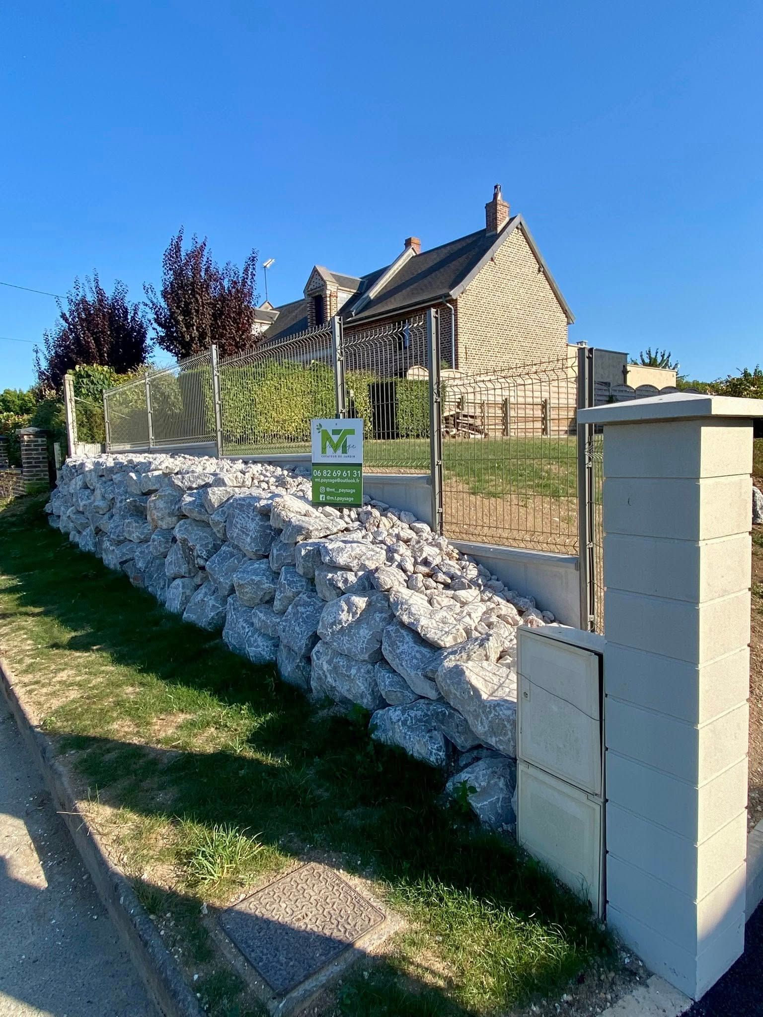 Un mur de soutènement en pierre, une pente avec de l'herbe et une clôture en fil de fer devant une maison en pierre sous un ciel bleu limpide.