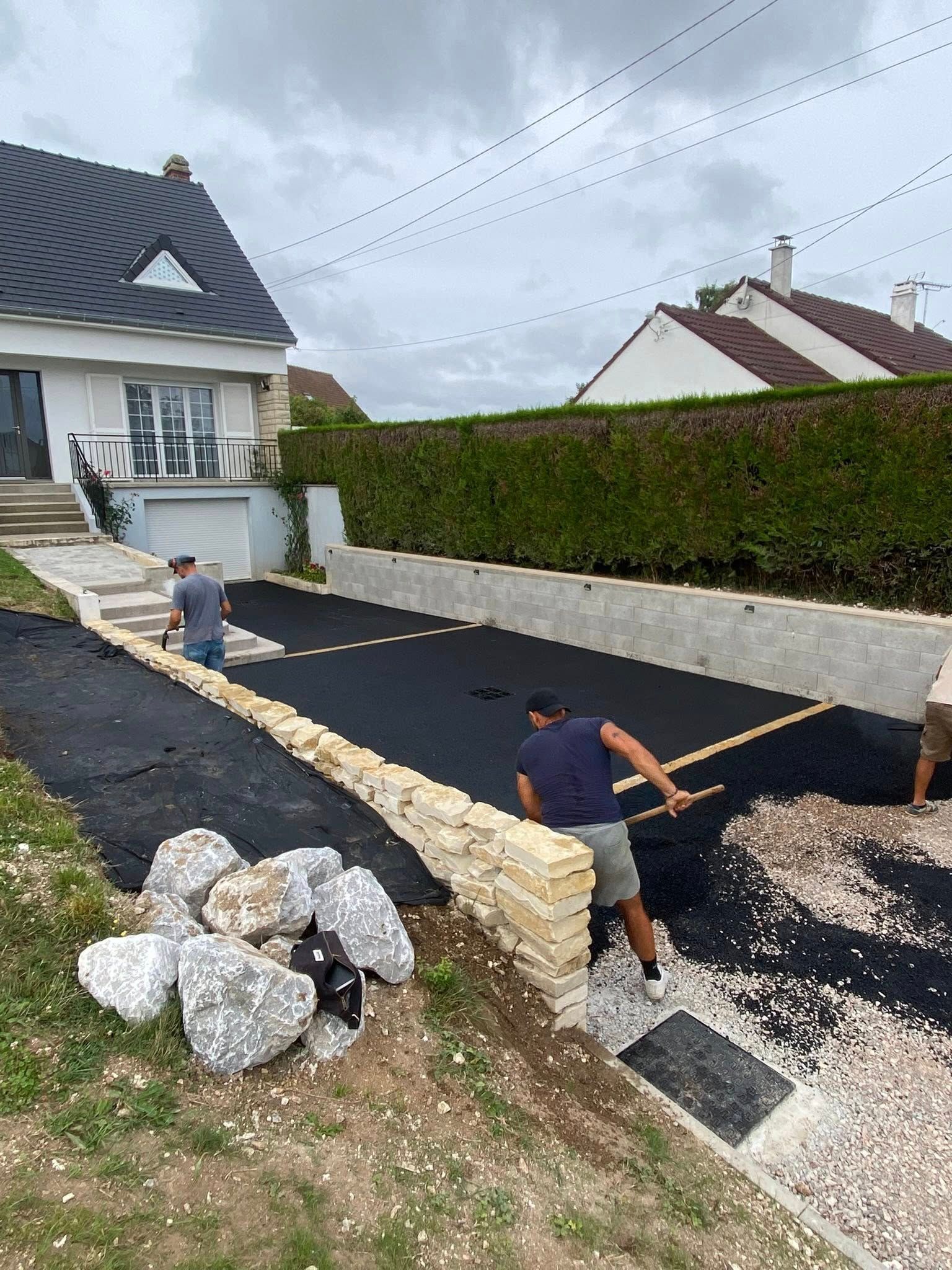 Deux personnes installent une toile de paillage noire sur une allée de gravier, à côté d'un mur de soutènement en pierre nouvellement construit.